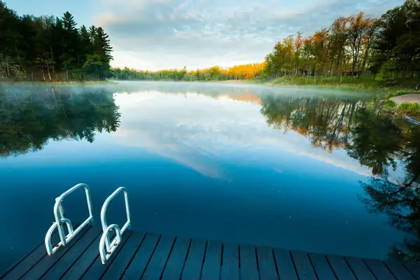 A dock with a ladder over a still lake surrounded by trees and reflecting the sky