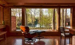 Interior view of a room with wooden walls large windows overlooking trees and a lake featuring a lounge chair