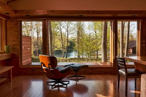Interior view of a room with wooden walls large windows overlooking trees and a lake featuring a lounge chair