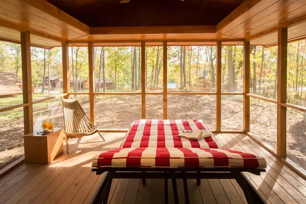 A cabin room with a striped bed a chair and surrounding windows showing an outdoor wooded view