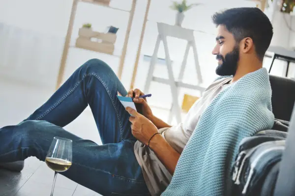 young man writing a letter hes sitting on the floor, drinking white wine and thinking about his loved one while hes writing her a love letter