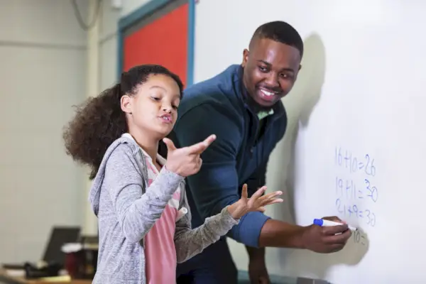 a multiracial 9 year old girl in math class, standing at the whiteboard with her teacher, a young african american man in his 20s the teacher is looking at his elementary student, waiting while she tries to figure out the answer the focus is on the girl