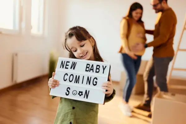 happy little girl holding announcement message with a text that new baby is coming soon with her parents in the background