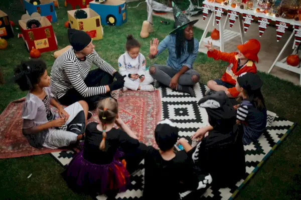 parents telling scary stories to kids at outdoor halloween party they are sitting cross legged on rugs on the grass, one parent is in a witch