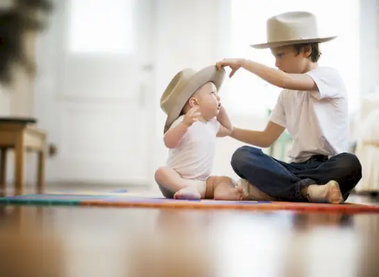 selective focus view of two brothers, one is nine one is eight months the pair sit cross legged on the floor older boy puts matching hat on babys head