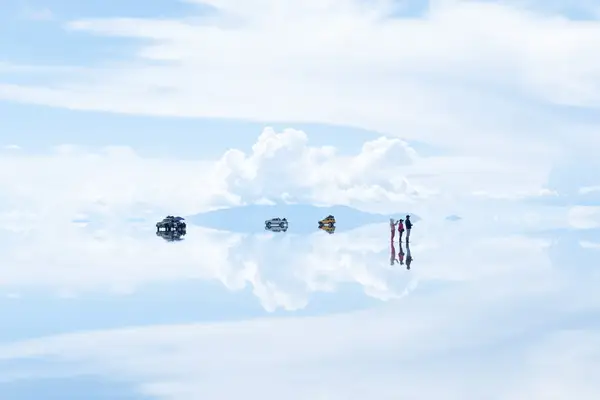Reflection of vehicles and people on a salt flat under a vast sky with clouds