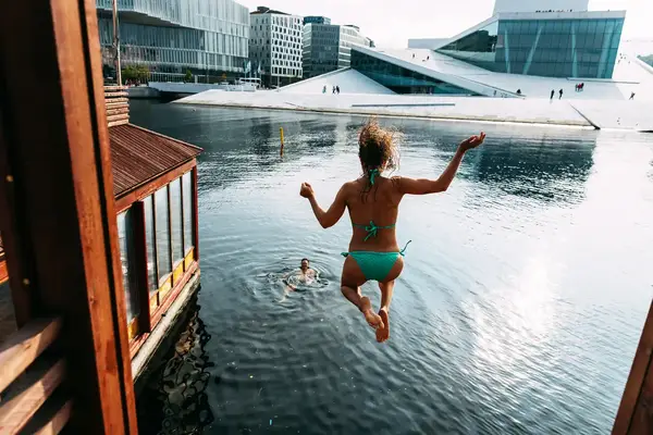 Person jumping into water near a city waterfront another person swimming below