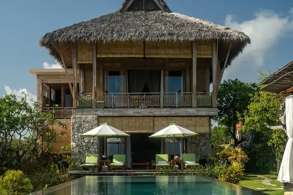 A twostory tropical villa with a straw roof overlooking a swimming pool surrounded by lush greenery