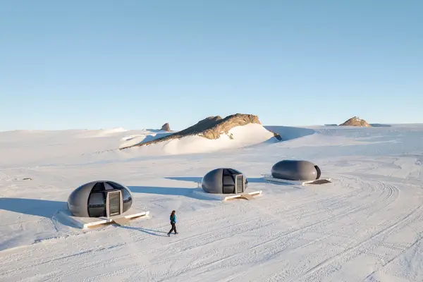 Three futuristic pods on a snowy landscape with a person walking nearby Antarctic setting with rocky outcrops in the distance