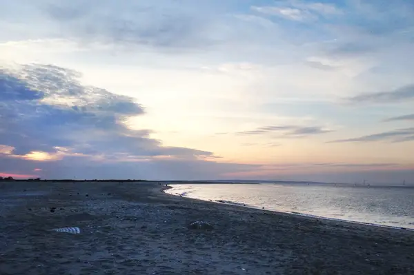 A sandy beach at sunset, with calm ocean waves and a partly cloudy sky in the background