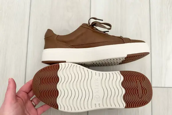 A hand showing the bottom of one Kizik Women’s Vegas Sneakers on a wooden floor