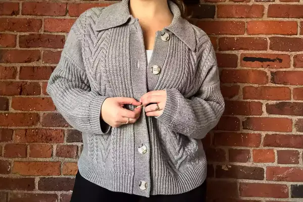 A person wearing the Quince Australian Merino Wool Collared Cable Cardigan standing in front of a brick wall