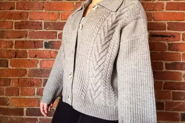 A person wearing the Quince Australian Merino Wool Collared Cable Cardigan standing in front of a brick wall