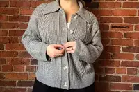 A person wearing the Quince Australian Merino Wool Collared Cable Cardigan standing in front of a brick wall