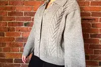 A person wearing the Quince Australian Merino Wool Collared Cable Cardigan standing in front of a brick wall