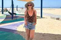 A person wearing a hat, sunglasses, andTA3 Brazilian Plungey Swimsuit, standing in a playground area on a sunny beach