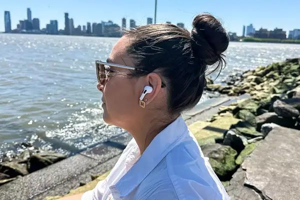 A person wearing sunglasses and Apple AirPods Pro 2 sits near a waterfront, with a city skyline in the background