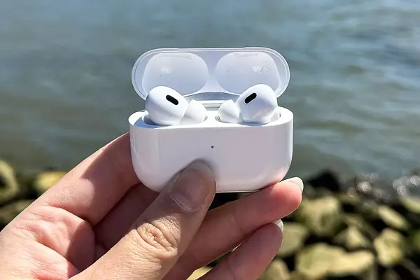 A hand holding a pair of Apple AirPods Pro 2 in an open case near a body of water