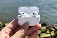 A hand holding a pair of Apple AirPods Pro 2 in an open case near a body of water