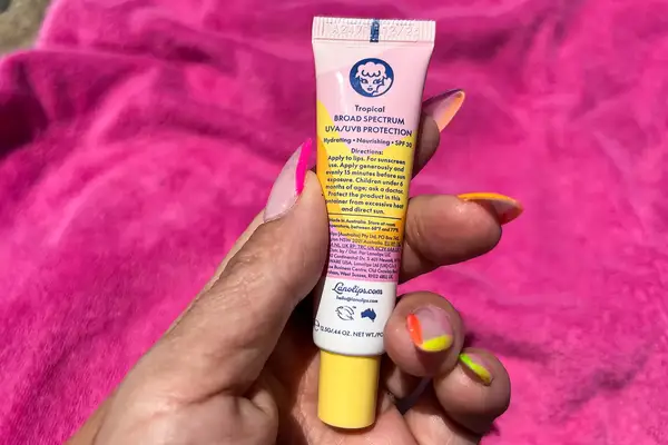 A tube of Lanolips Sun Balm SPF 30 held in a hand with colorful nails, a pink towel in the background