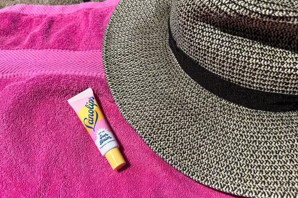 A tube of Sun Balm by Lanolips on a pink towel next to a wide-brimmed hat