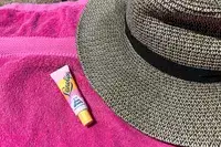 A tube of Sun Balm by Lanolips on a pink towel next to a wide-brimmed hat