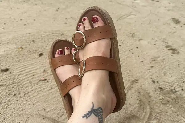 A person wearing the Beek Warbler Leather Platform Platform Leather Slides with their feet crossed and a sand background