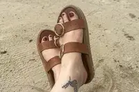 A person wearing the Beek Warbler Leather Platform Platform Leather Slides with their feet crossed and a sand background