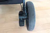 Closeup of a rubber wheel with a black plastic axle and pedal of Nuna TRVL LX Stroller on a wooden floor