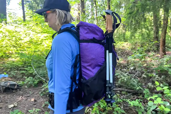 A person outdoors wearing a ULA Equipment Circuit with hiking poles attached, in a forested area