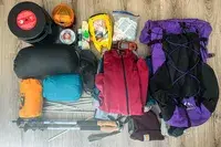 Assorted camping and hiking gear, including a ULA Equipment Circuit, clothing layers, a bear canister, and various small items, laid out on a floor