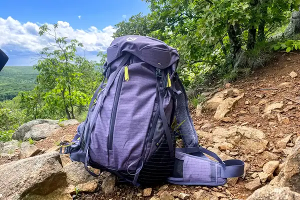 A NEMO Persist 45 L Endless Promise All-Adventure Pack rests on a rocky outdoor trail amidst greenery and trees