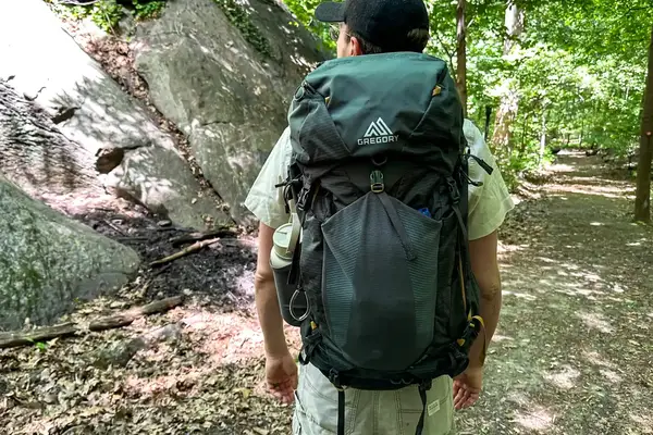 A hiker walking on a forest trail wearing a Gregory Katamai 55 - Hiking Backpack with camping gear, nature surrounding the path