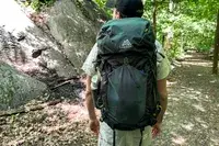 A hiker walking on a forest trail wearing a Gregory Katamai 55 - Hiking Backpack with camping gear, nature surrounding the path