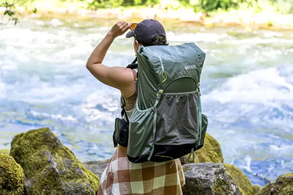 A person wearing a Gossamer Gear Mariposa 60 Backpack stands near a river, holding sunglasses and looking into the distance