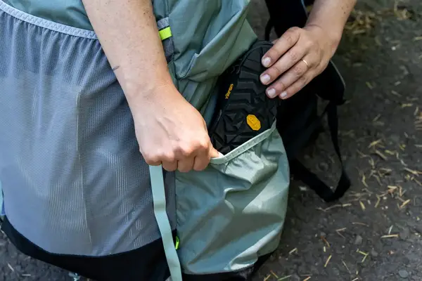 A person placing footwear into the side pocket of a Gossamer Gear Mariposa 60 Backpack outdoors