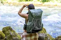 A person wearing a Gossamer Gear Mariposa 60 Backpack stands near a river, holding sunglasses and looking into the distance