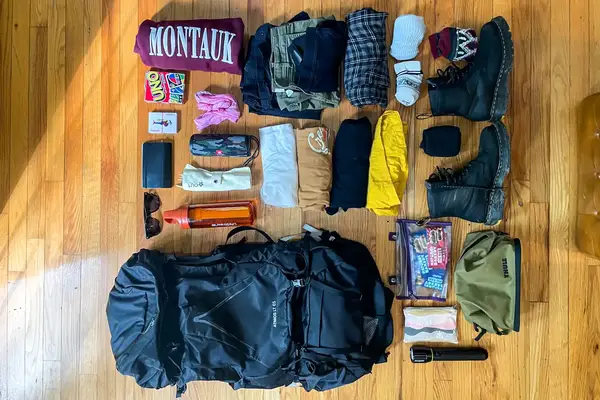 Assorted travel gear laid out on a wooden floor, including clothing, a Osprey Atmos AG 65 Men