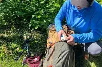 A person in outdoor clothing organizing items in a The North Face Trail Lite 50 Backpack on the ground near trekking poles and other outdoor gear, surrounded by greenery