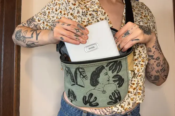 A person placing a white Instax device into a fanny pack with an artistic design showing detailed tattoos on their arms and hands