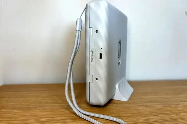 A white electronic device with a wrist strap resting on a wooden surface viewed from the side showing ports and branding