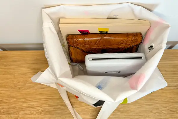 A fabric tote bag containing a book with sticky notes a leather wallet and a white electronic device placed on a wooden surface