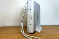 A white electronic device with a wrist strap resting on a wooden surface viewed from the side showing ports and branding