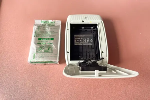 An Instax Mini film packet next to an open Instax Mini printer displaying its empty film compartment