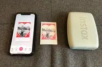 A smartphone displaying a photo editing app a printed photo with a red heart border and an Instax mini printer on a gray surface