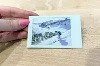 A hand holding a photo of a skier descending a snowy slope with buildings and mountains in the background