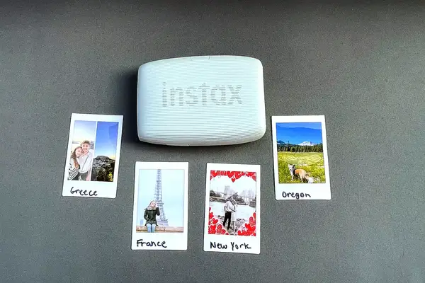 A white Instax mini printer surrounded by four printed photographs labeled with locations Greece France New York and Oregon