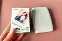 A hand holding a box of Fujifilm Instax Mini film packets next to a portable photo printer