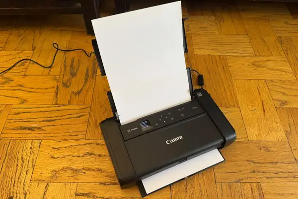 A Canon PIXMA TR150 wireless portable printer on a wooden floor with paper loaded for printing