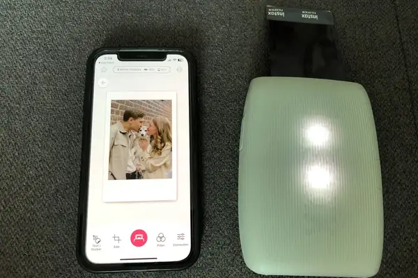 A smartphone displaying an edited photo of a couple with a dog alongside a Fujifilm Instax mini printer